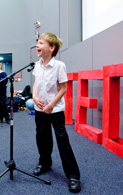 A young boy in a white shirt and black pants stands at a microphone, smiling as he speaks. Behind him are large red letters spelling TED on a stage. The setting appears to be a conference or presentation event.