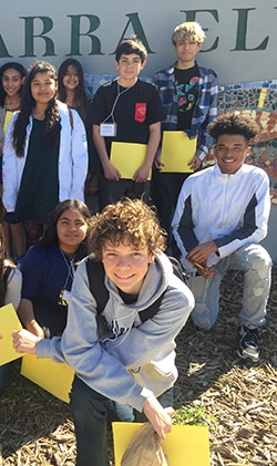 A group of students posing outdoors in front of a mural. Some are holding yellow papers. They are casually dressed, with a mix of T-shirts, hoodies, and jackets. Smiles and sunlight create a cheerful atmosphere.