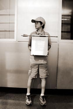 A young boy wearing a cap and casual clothes stands holding a framed document. The background shows a wall with windows.