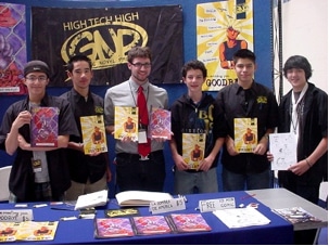 A group of six young men stand behind a table displaying comic books and illustrations. They are holding up various comic issues. A banner above them reads High Tech High. The scene appears to be at a comic convention or school event.