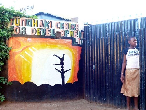 A person stands beside a colorful mural on a wall that reads Tunohaki Centre for Development. The mural features a silhouette of a dancer against a vibrant sun-like background. The person is wearing a t-shirt and a skirt, looking to the side.