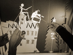 Hands manipulate shadow puppets of a stick figure and a horse over a cityscape backdrop with a tree and star. The scene is monochrome, with a creative and whimsical atmosphere.