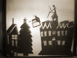 Silhouette of a winter scene with a reindeer and sleigh flying over a town. A stick-figure person rides the sleigh. Buildings and a large Christmas tree with a star on top are visible in the foreground. Vintage sepia tone.