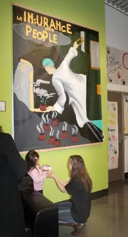 A child is seated on a couch while a woman kneels in front of her holding her hands. They are in a room with a large mural on the wall depicting a person in a lab coat pouring coffee from a test tube titled La Insurance People.