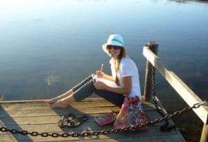 A person sitting on a wooden dock by a lake, smiling and wearing a sun hat and sunglasses, with a notepad in hand. A colorful bag and sandals are nearby, with calm water in the background under a clear sky.