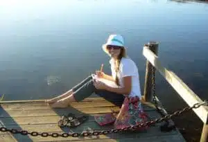 A person sitting on a wooden dock by a lake, smiling and wearing a sun hat and sunglasses, with a notepad in hand. A colorful bag and sandals are nearby, with calm water in the background under a clear sky.