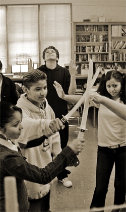 A group of young people in a classroom gather in a circle, each holding a sword with the tips meeting in the center. Bookshelves and large windows are in the background. One person in the back stands with arms raised, looking upwards.
