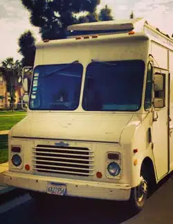 A white food truck with a classic design is parked on a street. The front of the truck has a large windshield and a license plate is visible. Trees and a grassy area are in the background under a partly cloudy sky.