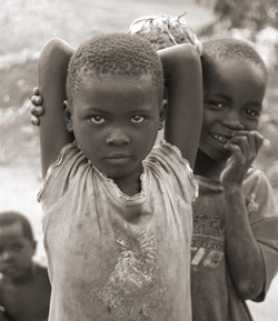 A sepia-toned image shows three children. The child in front has their arms crossed above their head and looks directly at the camera. Another child behind them is smiling and biting their finger. The background is blurred.