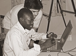 Two people sit closely at a table, focused on a laptop. The person in front is typing, while the other leans over to assist. Both are engaged in a collaborative activity, with a camera tripod visible in the background. The image is in black and white.