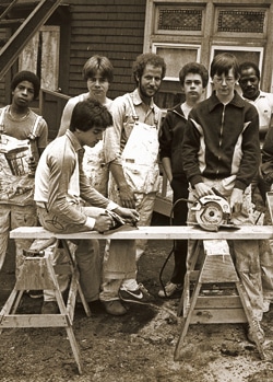A group of people, some in work overalls, gather around a wooden workbench outdoors. A man in front is operating a power saw on a piece of wood. The building behind has detailed windows and a staircase. The photo is in black and white.