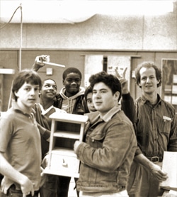 A group of young adults stand together in a room, one holding a small wooden object. They smile and pose casually, with one person making a peace sign. The room has visible bulletin boards and shelves in the background.
