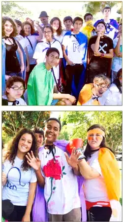 A group of people outdoors, dressed in colorful superhero capes and shirts with symbols. They are smiling and waving, suggesting a fun or themed event. Trees and blue sky are visible in the background.