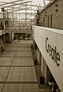 Interior view of a modern building with a high glass roof and brick walls. The foreground has a large sign that reads Create. The floor is tiled, and there are stairs and glass doors at the back. The sepia tone adds a vintage feel.