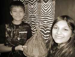Two children smiling and holding a small model structure made of sticks. The background features a zebra print pattern. The image is in black and white.