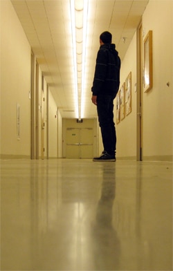 A person in a dark sweater stands in a long, brightly lit hallway with closed doors. The floor reflects the overhead lights, and artwork hangs on the walls. The person faces away from the camera, towards a set of double doors at the end.