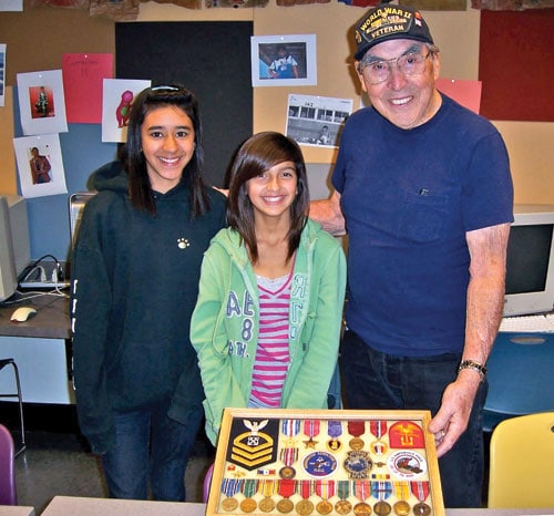 A veteran wearing a cap stands smiling with two young girls in a classroom. He holds a display case filled with various medals and insignia. The group is standing near colorful chairs and walls decorated with photos.