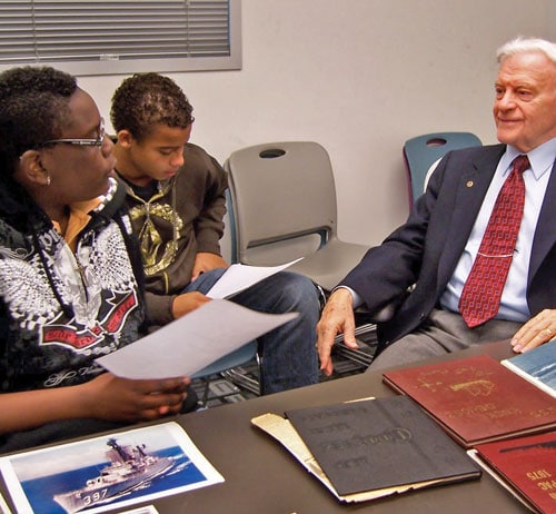 A man in a suit sits at a table with two people. They are looking at documents and a photo of a ship. The room has a casual setting with chairs and a window in the background.