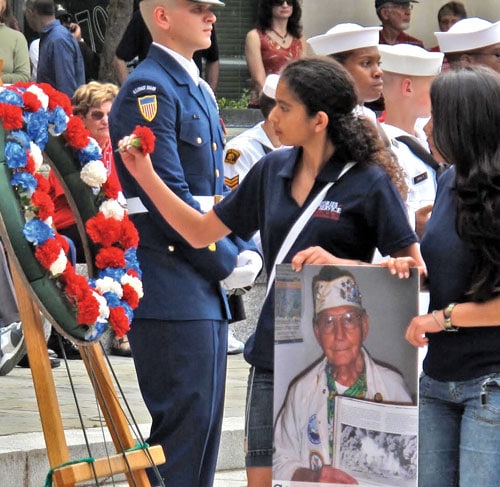 A young woman places a flower on a wreath during a memorial ceremony. She holds a photograph of an elderly veteran. Military personnel in uniform stand solemnly in the background.
