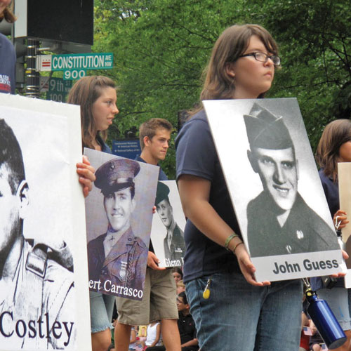 A group of people at a march hold up large black-and-white photos of military personnel. They are walking along a street with lush green trees in the background. Signs are visible, including Constitution Ave and 900.