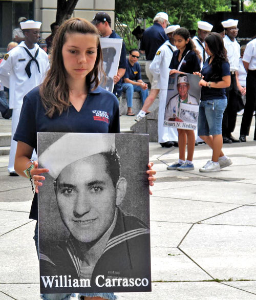 A young woman holds a poster of a sailor named William Carrasco, honoring him in a ceremony. In the background, participants in navy uniforms and civilians hold similar posters.