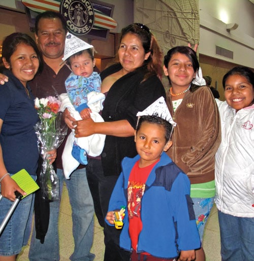 A family of seven, including two adults, four children, and a baby, pose happily indoors. Some children wear paper hats, one holds a toy car, and the woman holds flowers. They stand in front of a coffee shop sign, smiling at the camera.