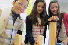 Three children are working together with wooden blocks in a classroom setting as part of an engaging educational campaign. One child wears safety goggles, and the other two smile as they interact, surrounded by colorful notes on the wall, highlighting this creative learning medium.