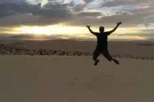 A person jumps joyfully in a desert landscape at sunset, silhouetted against a colorful sky with scattered clouds. The vast sand dunes stretch into the distance under the fading light.