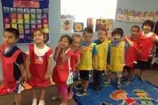 A group of eight children standing in a line indoors, wearing colorful vests and name tags. They are in a classroom setting with educational posters and a colorful floor mat featuring a cartoon design. The children appear to be smiling and excited.