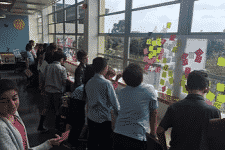 A group of children stands by large windows covered with colorful sticky notes, discussing and observing the view outside. The room is bright, with natural light streaming in, creating a lively backdrop perfect for an event listing photo that would captivate any sidebar browser.