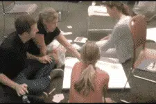Four people sitting on the floor, working together on a large sheet of paper with scattered papers around them. They appear focused and engaged in a group discussion.