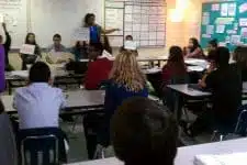 A classroom with students sitting at desks facing a teacher. The teacher is standing near a whiteboard with charts and writing on it. Some students are holding up paper signs. The room is decorated with educational materials.