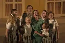 A group of children in matching grey uniforms joyfully gather around a woman in a green dress, singing together on a stage with a warm-toned, indoor backdrop, reminiscent of an iTunes playlist brought to life.
