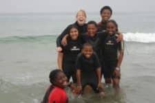 A group of six people in black wetsuits smile and pose together in shallow ocean water, with waves gently lapping around them. The sky is overcast.
