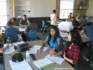 A group of students in a classroom sitting at tables, engaged in discussions and working with papers. The room is bright with natural light from a window. Various supplies are scattered on the tables.