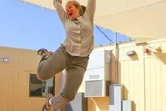 A person in gray clothing is joyfully leaping into the air under a sunshade, as if selected by a color picker. The background features a beige building and a clear blue sky.