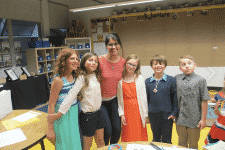 A group of children stand with an adult in a classroom. The children are smiling, and the adult, dressed in a red top, stands in the middle. Classroom decorations and shelves are visible in the background.