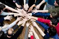 A diverse group of people standing in a circle, extending their hands towards the center over a wooden surface, symbolizing unity and teamwork.