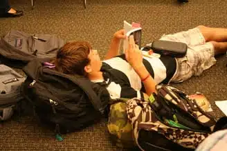 A person is lying on the floor reading a book, surrounded by backpacks. They are wearing a striped shirt and shorts, appearing relaxed and focused on their reading. The setting looks like an indoor carpeted area.