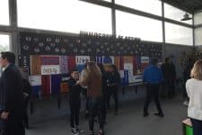 People stand in front of a wall displaying various flags and posters inside a building with large windows. The atmosphere appears busy and engaging.