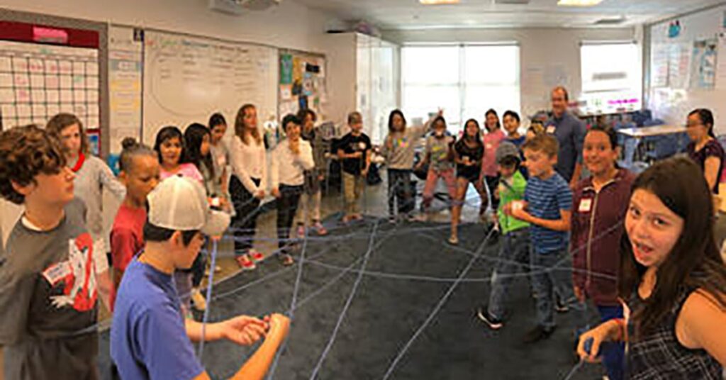 A group of children and adults stand in a classroom, forming a circle. They are connected by strings, creating a web pattern. The room has a whiteboard and several posters on the walls. Everyone is engaged and participating in an activity.