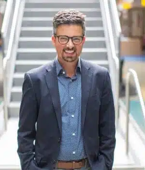 Man in a blue suit and polka-dot shirt stands smiling in front of a staircase. He has short dark hair, glasses, and a beard. The setting appears to be indoors, with blurred background details.