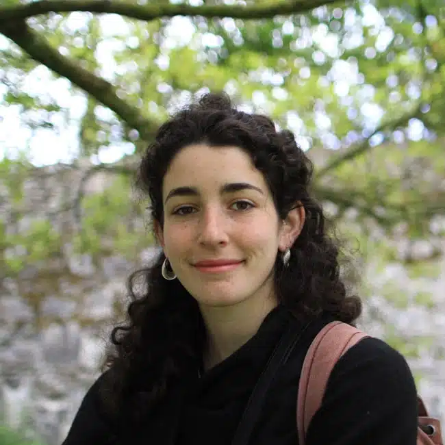 A person with curly hair smiles at the camera, standing outdoors in front of a backdrop of green trees and branches. They are wearing a black jacket and a backpack.