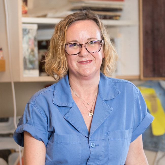 Amber George, a woman with short blonde hair and glasses, wearing a blue button-up shirt, smiles while standing indoors in a room with shelves and various items in the background.