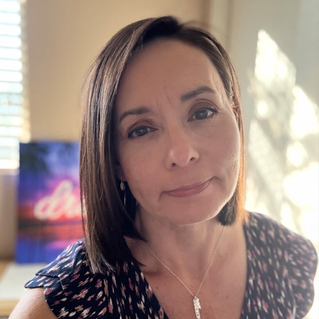 Anna Champion, a woman with straight, shoulder-length brown hair and a printed top, looks at the camera with a slight smile. She wears a necklace as sunlight filters into the softly lit room behind her.
