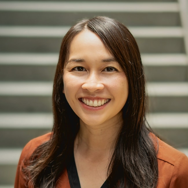 Smiling woman with long brown hair stands in front of a staircase, wearing a brown jacket and black top.
