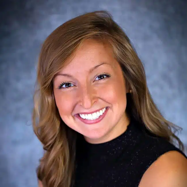 A woman with long, wavy hair smiles brightly. She wears a sleeveless black top, and the background is a textured gray.