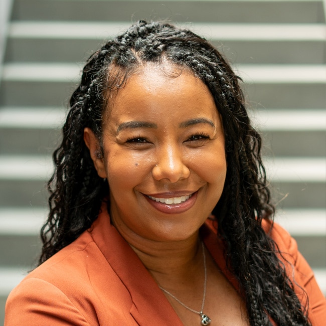 A woman with long, curly hair smiles warmly at the camera. She is wearing an orange blazer and a necklace. The background features blurred gray steps.