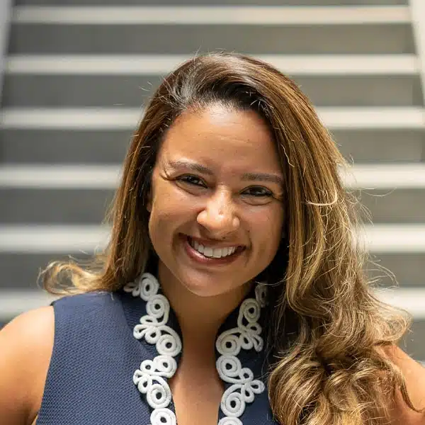 A person with long hair is smiling in front of a set of stairs. They are wearing a navy top with white decorative trim. The background is softly blurred.