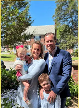 A family of four poses outdoors on a sunny day. The mother holds a young girl with a pink bow, while the father stands beside them. A young boy stands in front of them, smiling. They are surrounded by greenery and a brick house is visible in the background.
